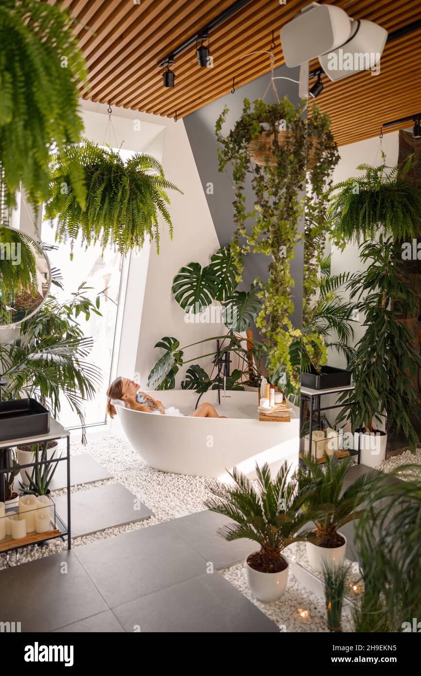 Relaxed young woman taking bubble bath in modern bathroom decorated with tropical plants at luxury spa resort Stock Photo