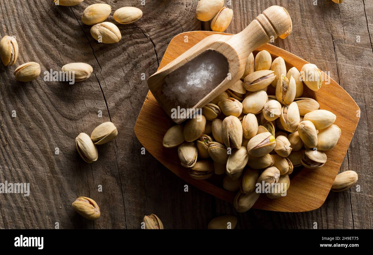 Heap of salted, roasted green pistachio nuts snack in wooden bowl on wood background with sea salt in wooden scoop, healthy food snack, selective focu Stock Photo
