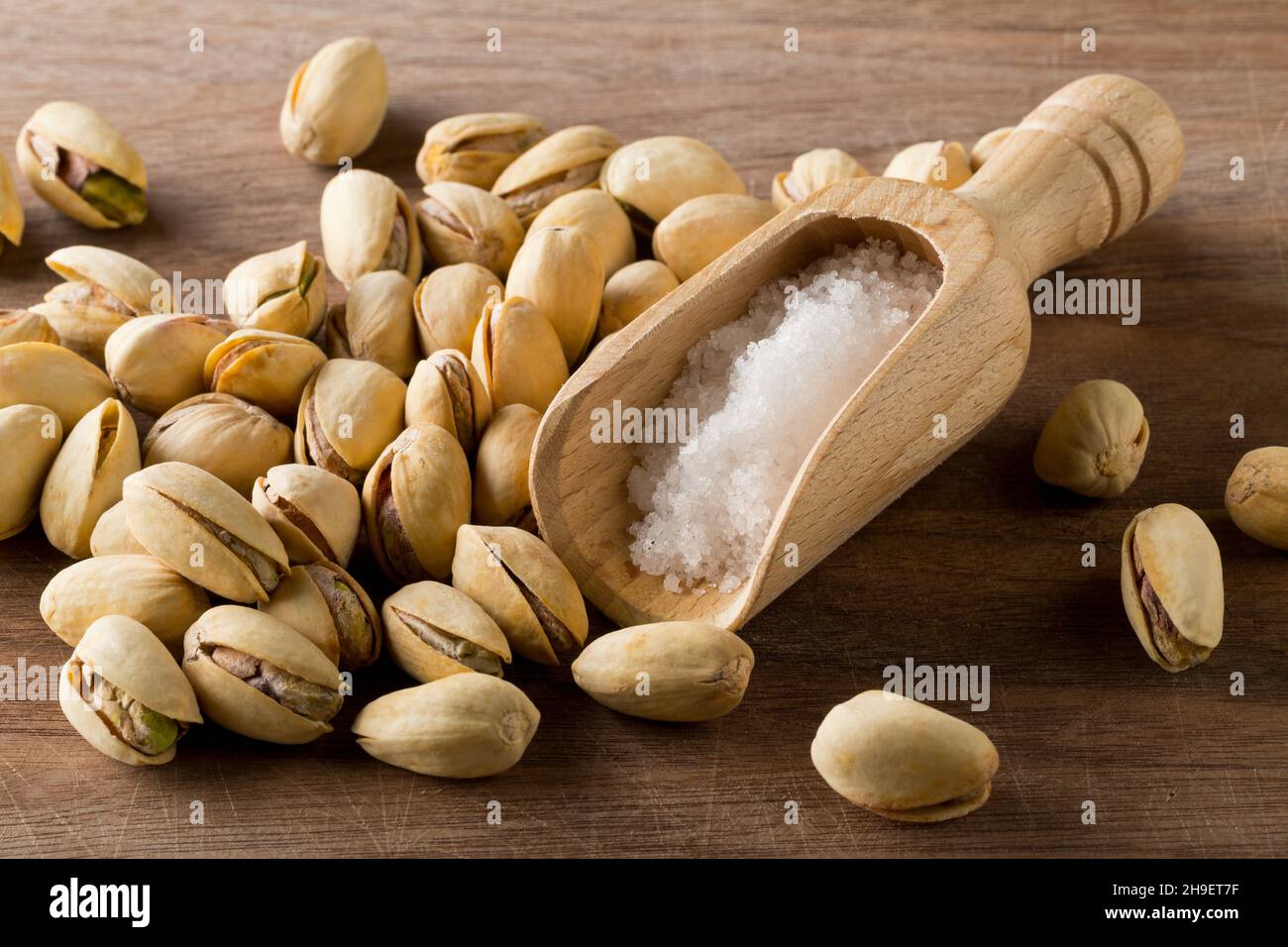 Heap of salted, roasted green pistachio nuts snack on wooden board background with sea salt in wooden scoop, healthy food snack, selective focus Stock Photo