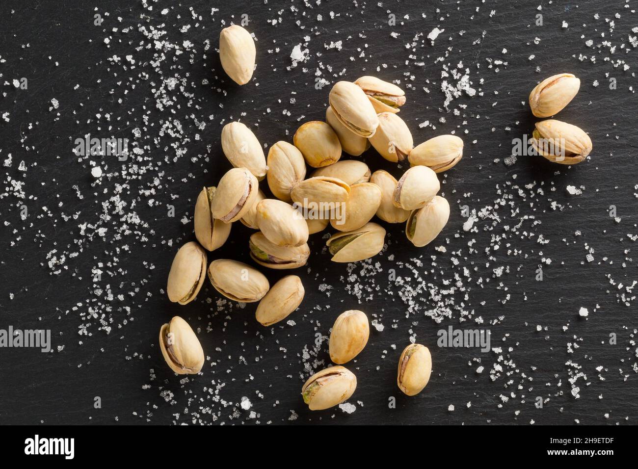 Heap of salted, roasted green pistachio nuts snack on black background with sea salt, healthy food snack, selective focus, flat lay top view from abov Stock Photo