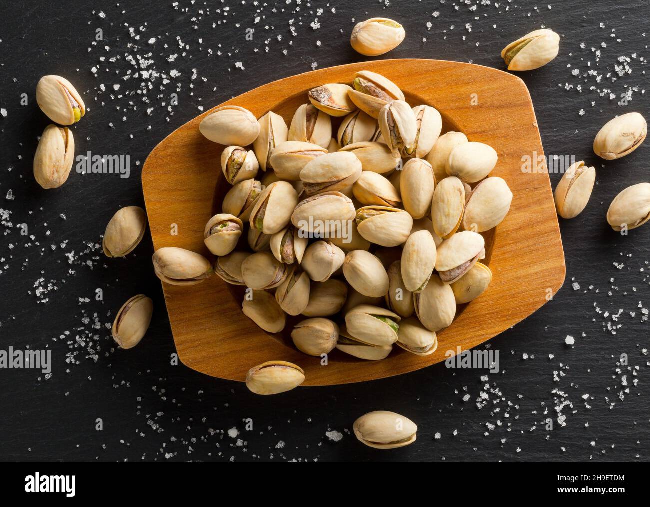 Heap of salted, roasted green pistachio nuts snack in wooden bowl on black background with sea salt, healthy food snack, selective focus, flat lay top Stock Photo