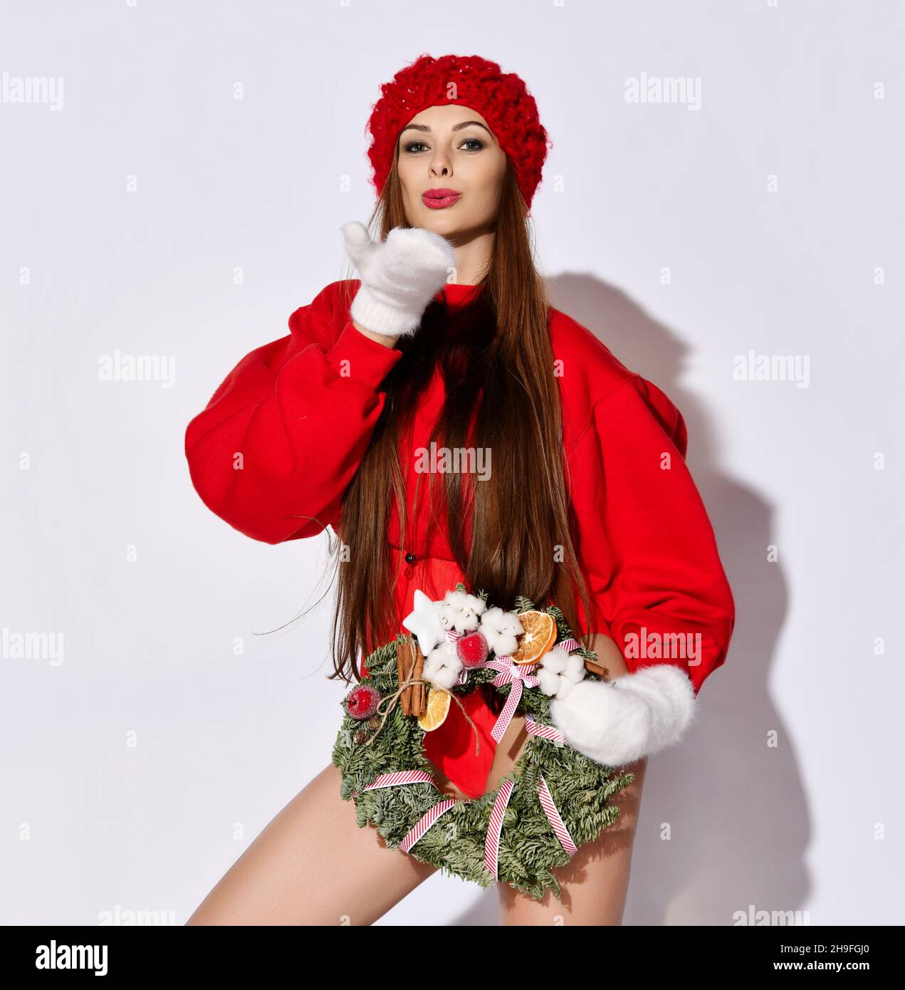 Cheerful girl in red short top sweater, bodysuit, hat and mittens with decorated Christmas wreath in hand sends a kiss Stock Photo