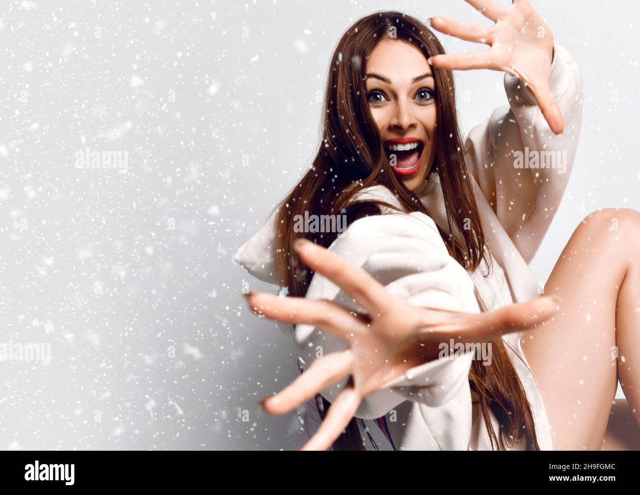 Cheerful excited woman with long hair in hoodie sits gesturing with hands and looking at camera under the snow Stock Photo