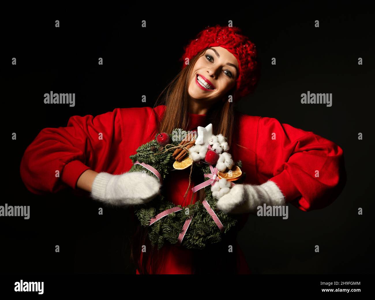 Happy cheerful woman in red winter sweater, warm knitted hat and mittens holds decorated Christmas ornate wreath  Stock Photo
