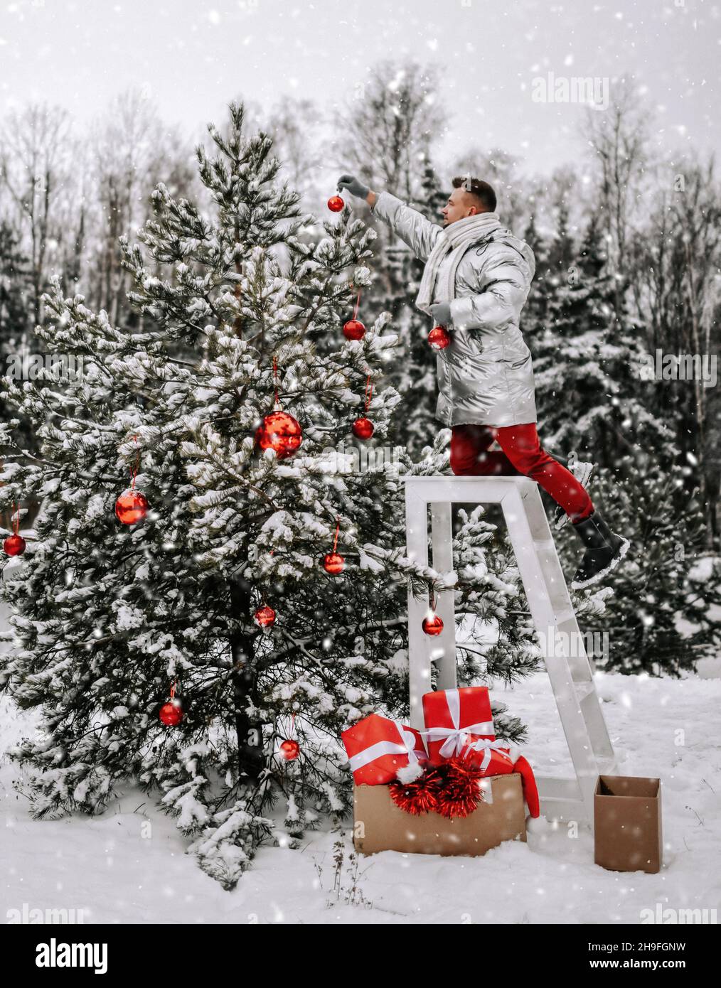 Young man guy in stylish winter clothing decorates Christmas tree with balls outdoors in winter snowy forest Stock Photo