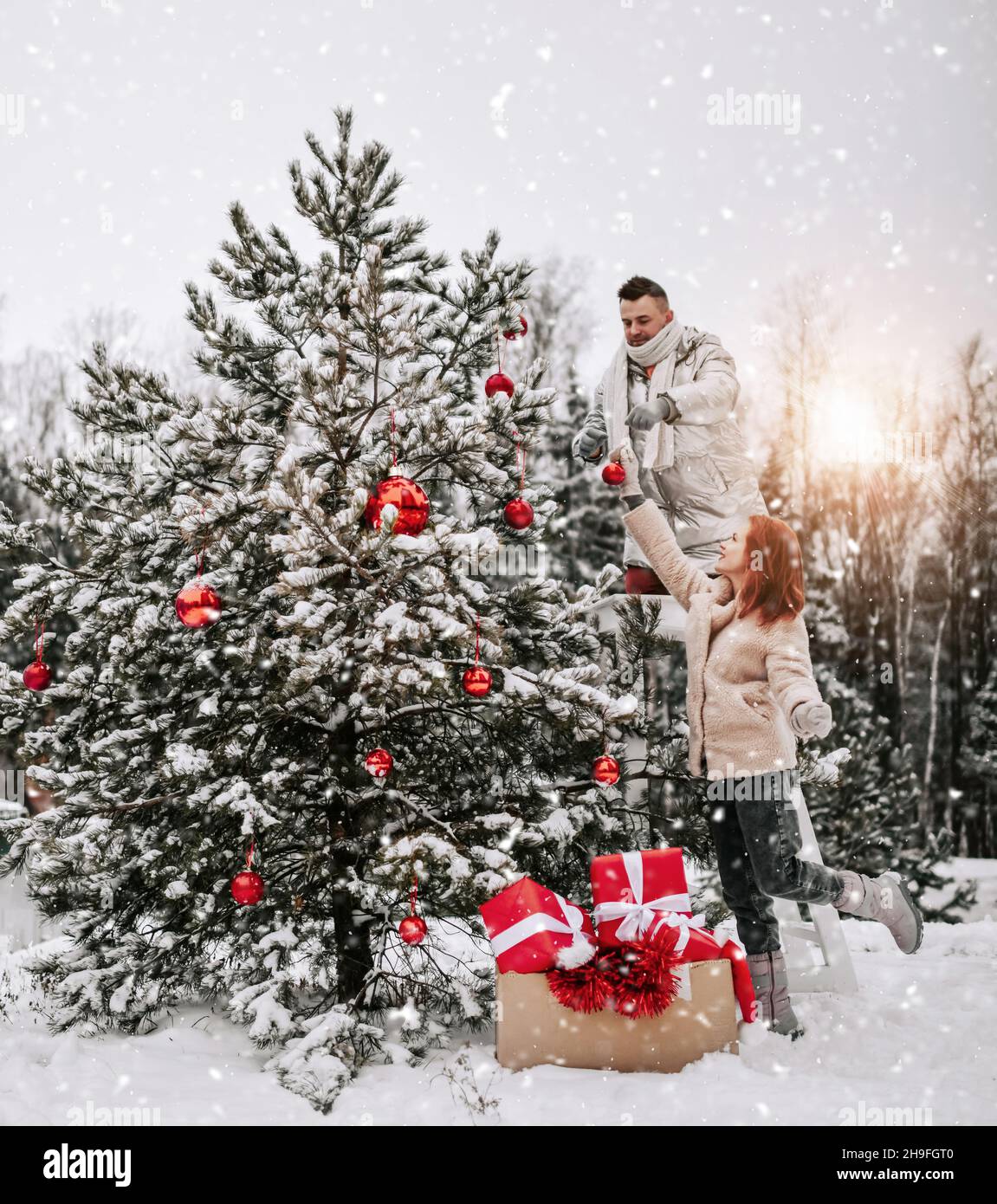 Young happy couple in stylish winter clothing decorating Christmas tree with red balls outdoors in winter snowy forest Stock Photo
