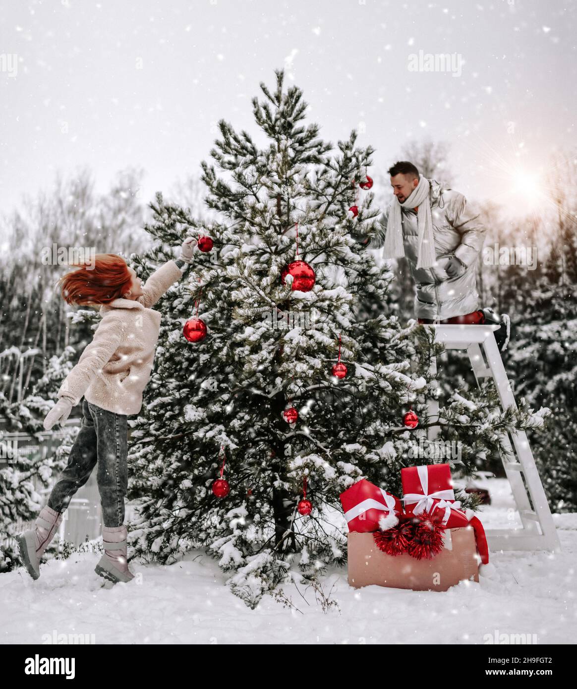 Young happy couple in stylish winter clothing decorating Christmas tree with balls outdoors in winter snowy forest Stock Photo