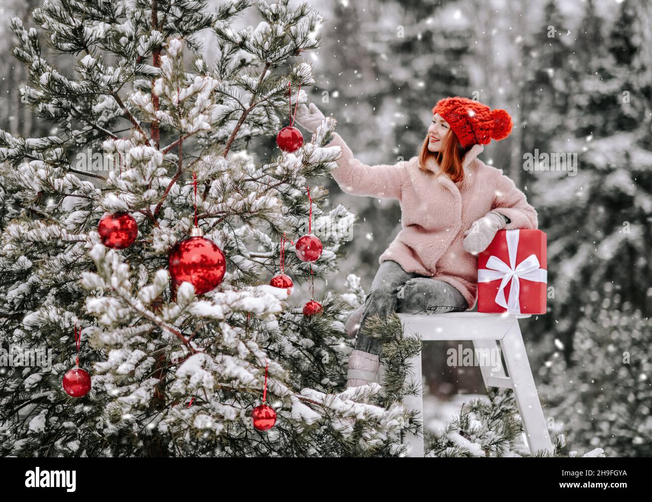 Young smiling pretty woman in stylish warm clothing sitting with giftbox on stepladder near decorated Christmas outdoors Stock Photo