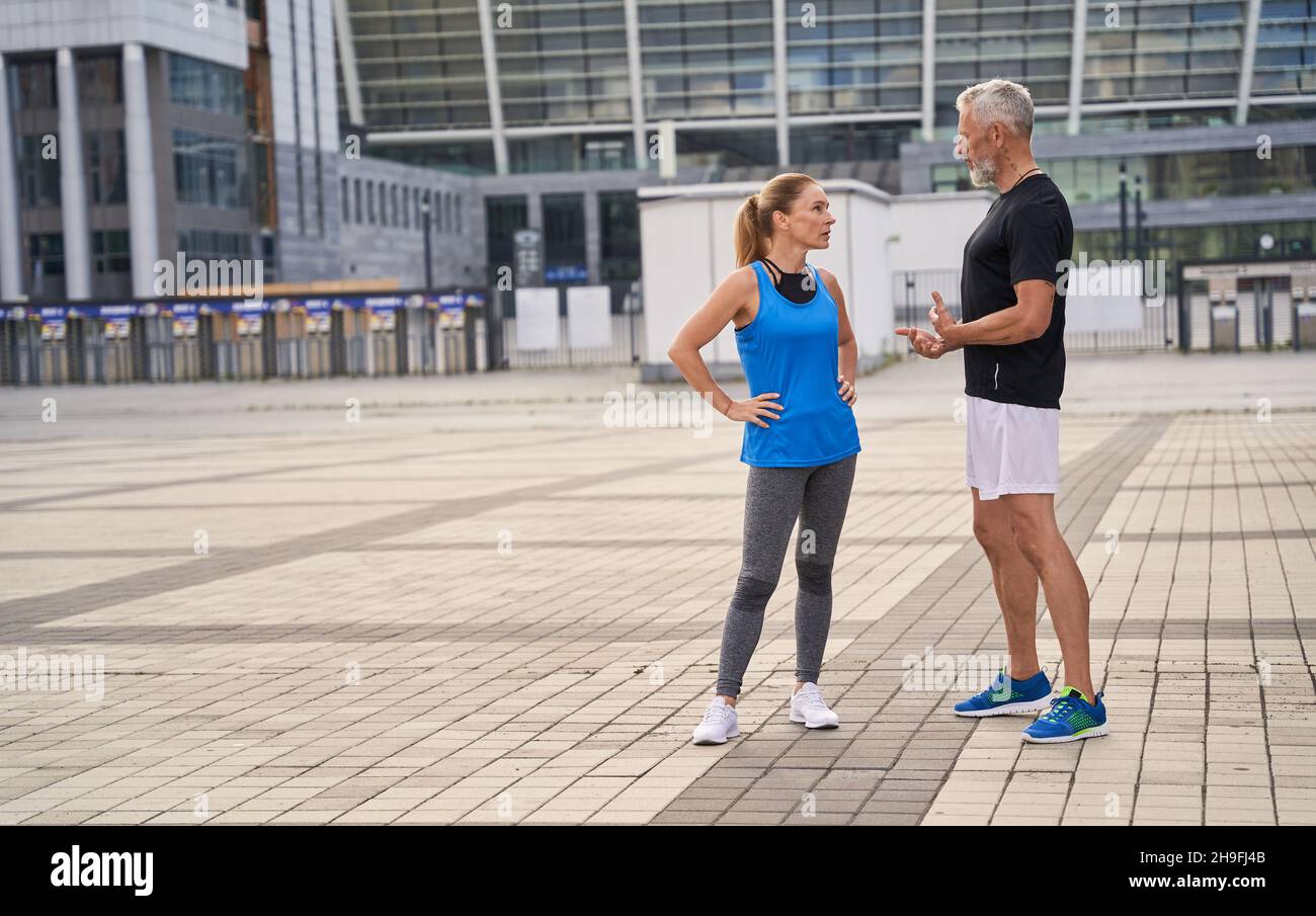Full length shot of two sportive friends, middle aged man and woman in sportswear talking while standing outdoors, doing sports together in the city Stock Photo