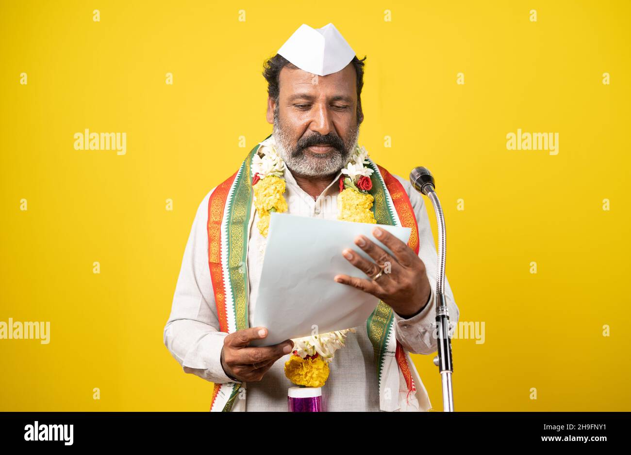 Politician seriously making oath ceremony with garland and white cap by holding papers in front of microphone on yellow background - concept of Stock Photo