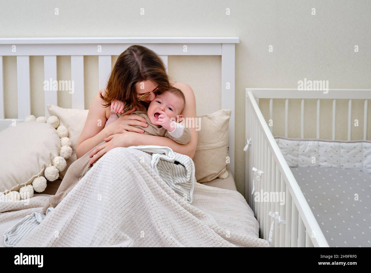A mother woman kisses and comforts a crying infant baby boy sitting on the bed Stock Photo