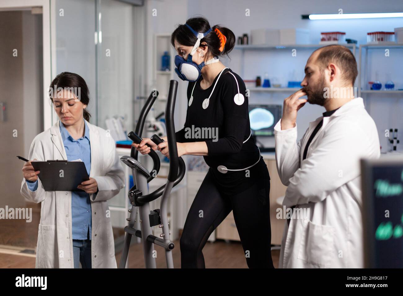 Athlete woman with mask running on gym bycle trainning body endurance while researcher doctor measuring heart rate monitoring EGK data in laboratory. Sportwoman with medical electrodes on it Stock Photo