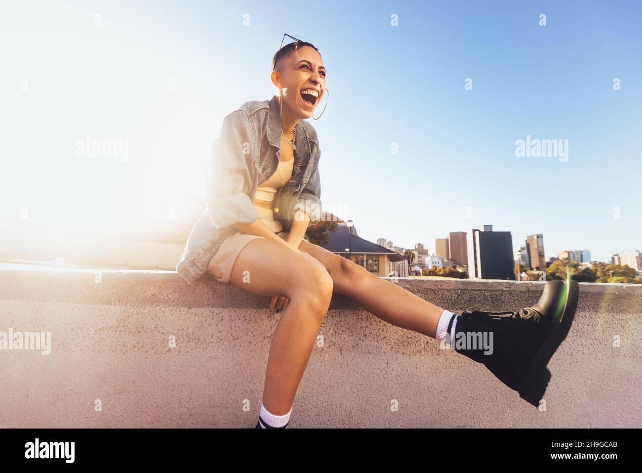 High-spirited young woman laughing cheerfully while sitting on a wall in the sun. Happy female youngster feeling vibrant and full of life in the city. Stock Photo