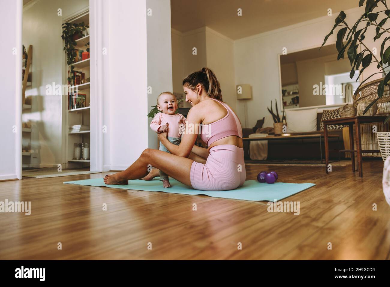 Young mother holding her adorable baby on an exercise mat. Happy mom working out with her little baby at home. New mom bonding with her baby during he Stock Photo