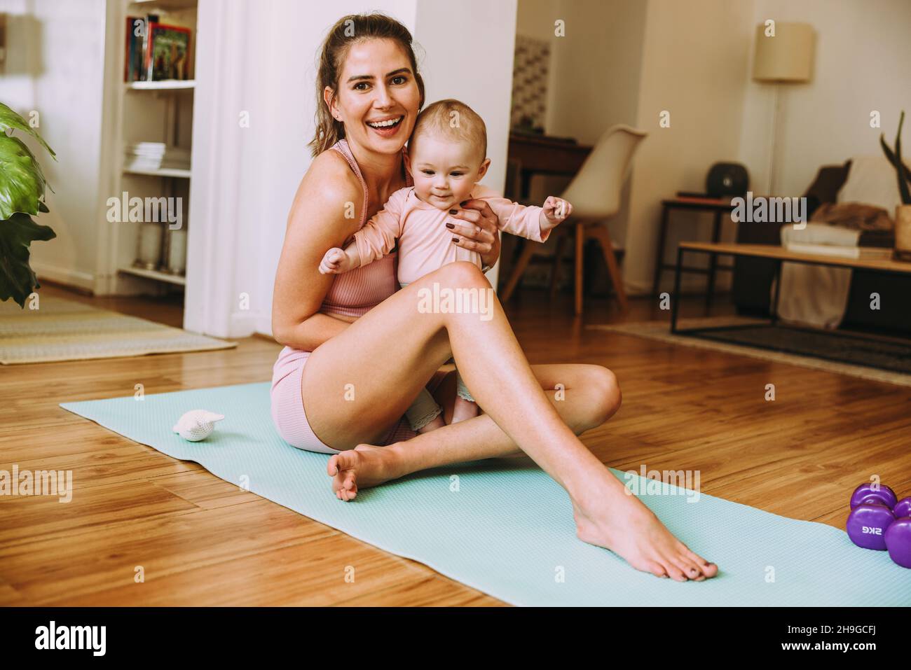 Smiling mom holding her baby while sitting on an exercise mat. Happy young mom working out with her little baby at home. New mom bonding with her baby Stock Photo