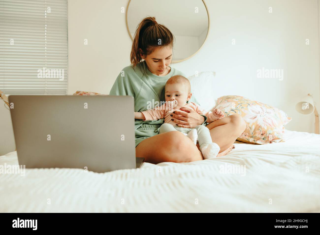 Student mom holding her baby while preparing for exams. Young single mother using a laptop to study while sitting on the bed with her baby. Academic m Stock Photo