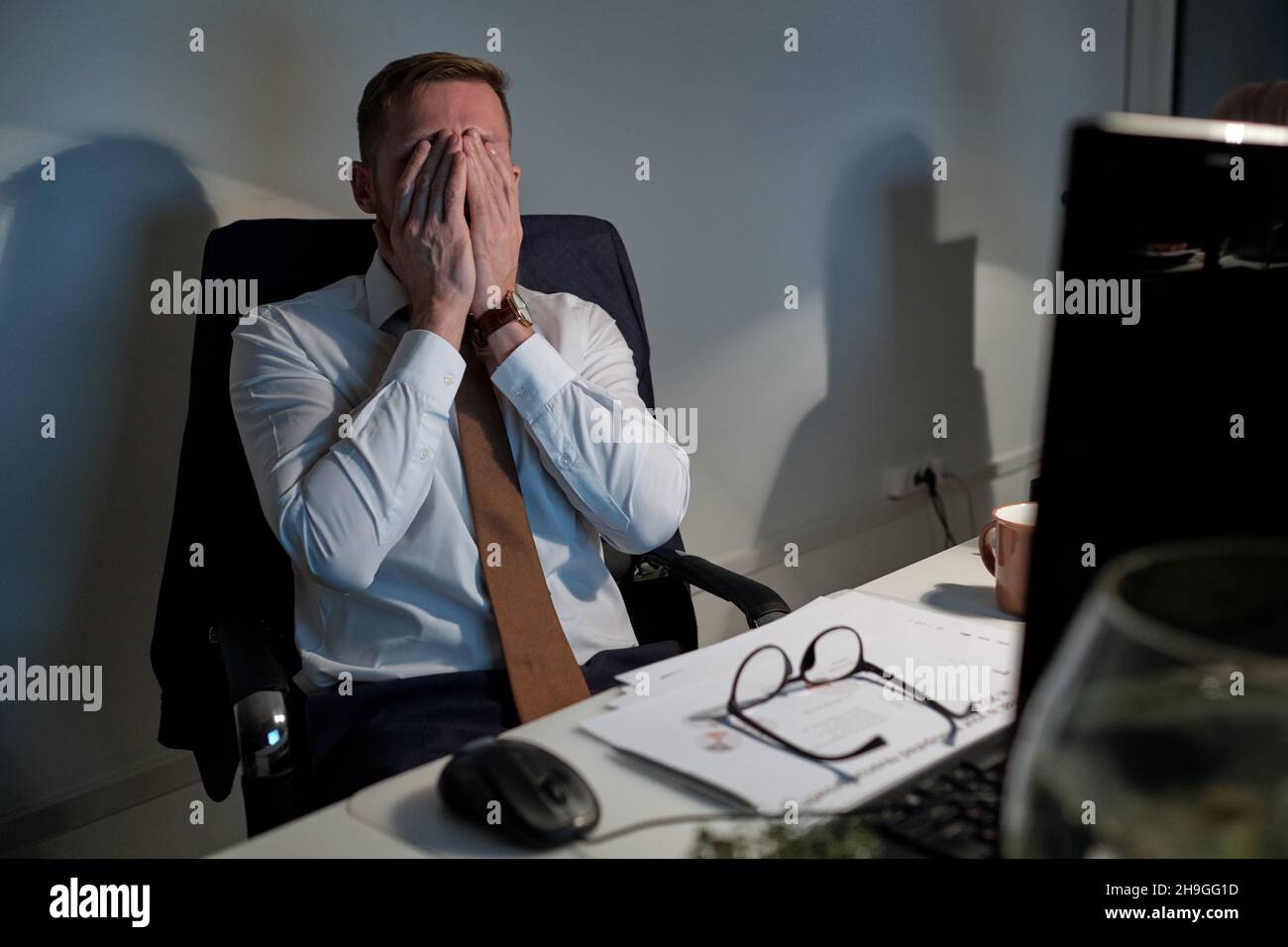 Exhausted businessman covering his face by hands while sitting in front ...