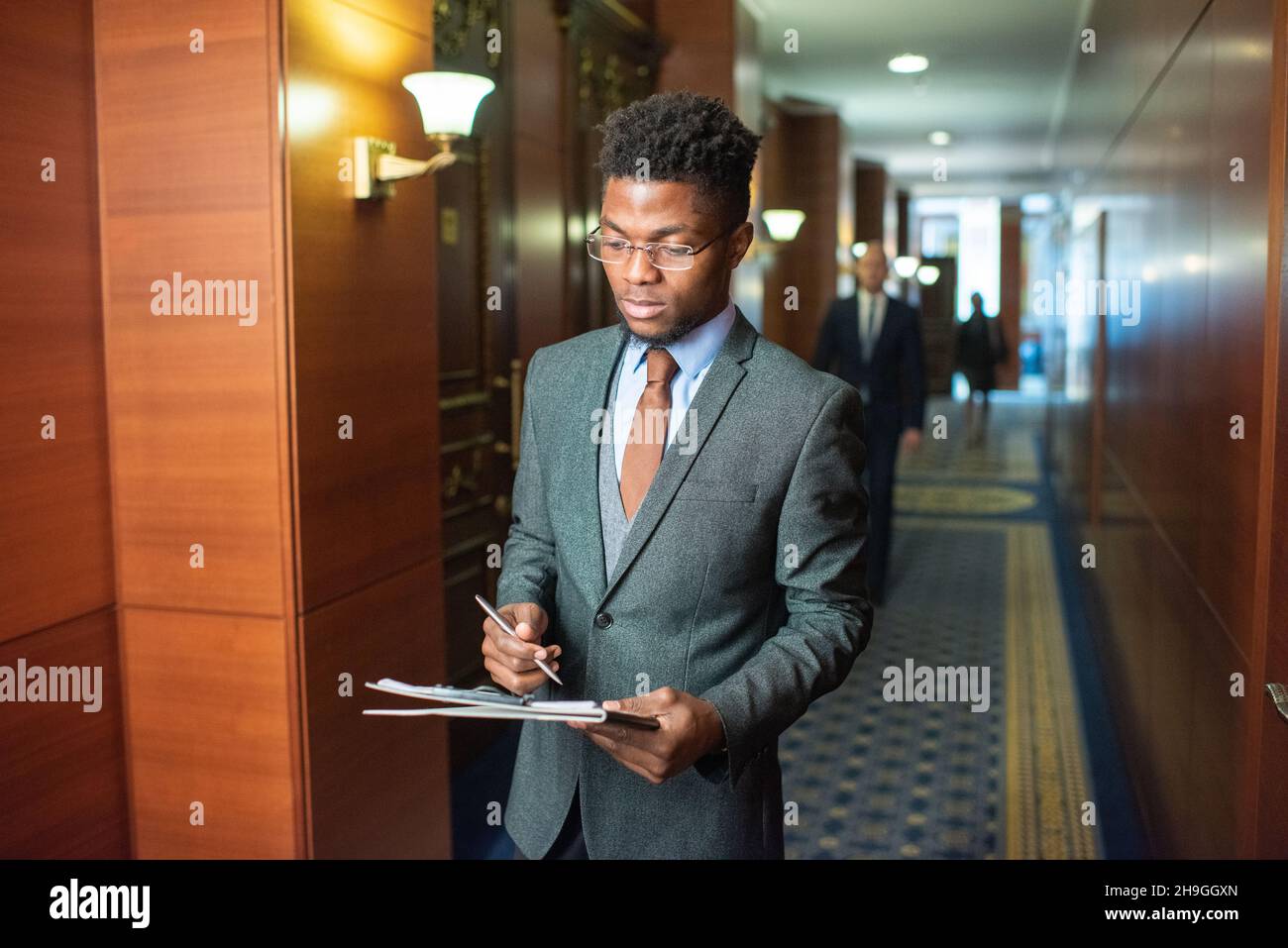 Young African businessman with clipboard standing in corridor of contemporary office center and reading document Stock Photo