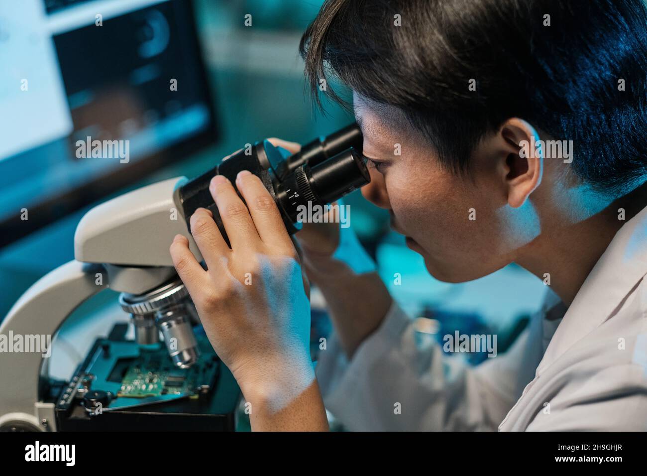 Young Asian technician looking in microscope while studying microchip characteristics in laboratory Stock Photo