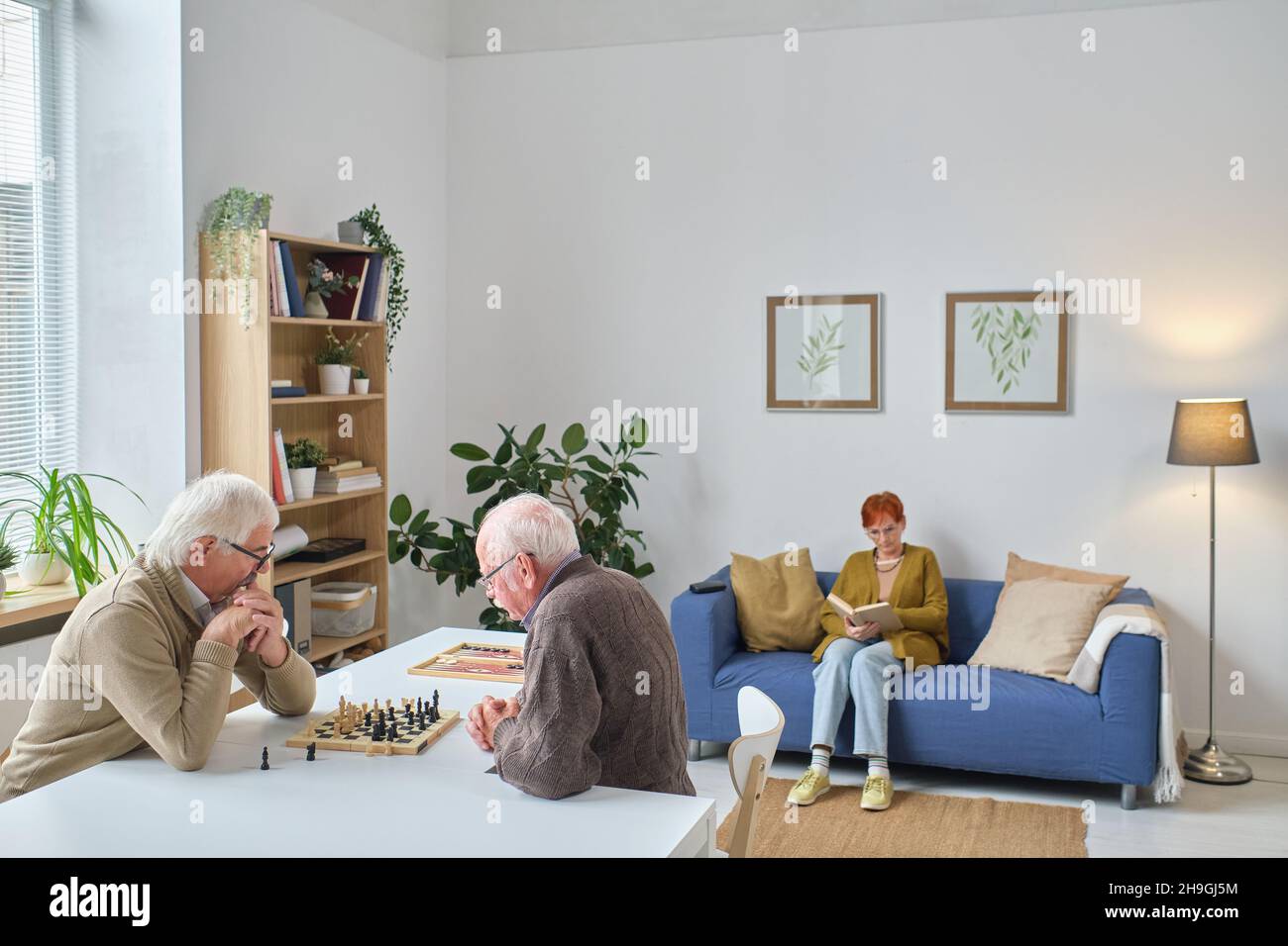 Two senior men sitting at the table and playing chess with woman reading a book on the sofa, they spending time in nursing home Stock Photo