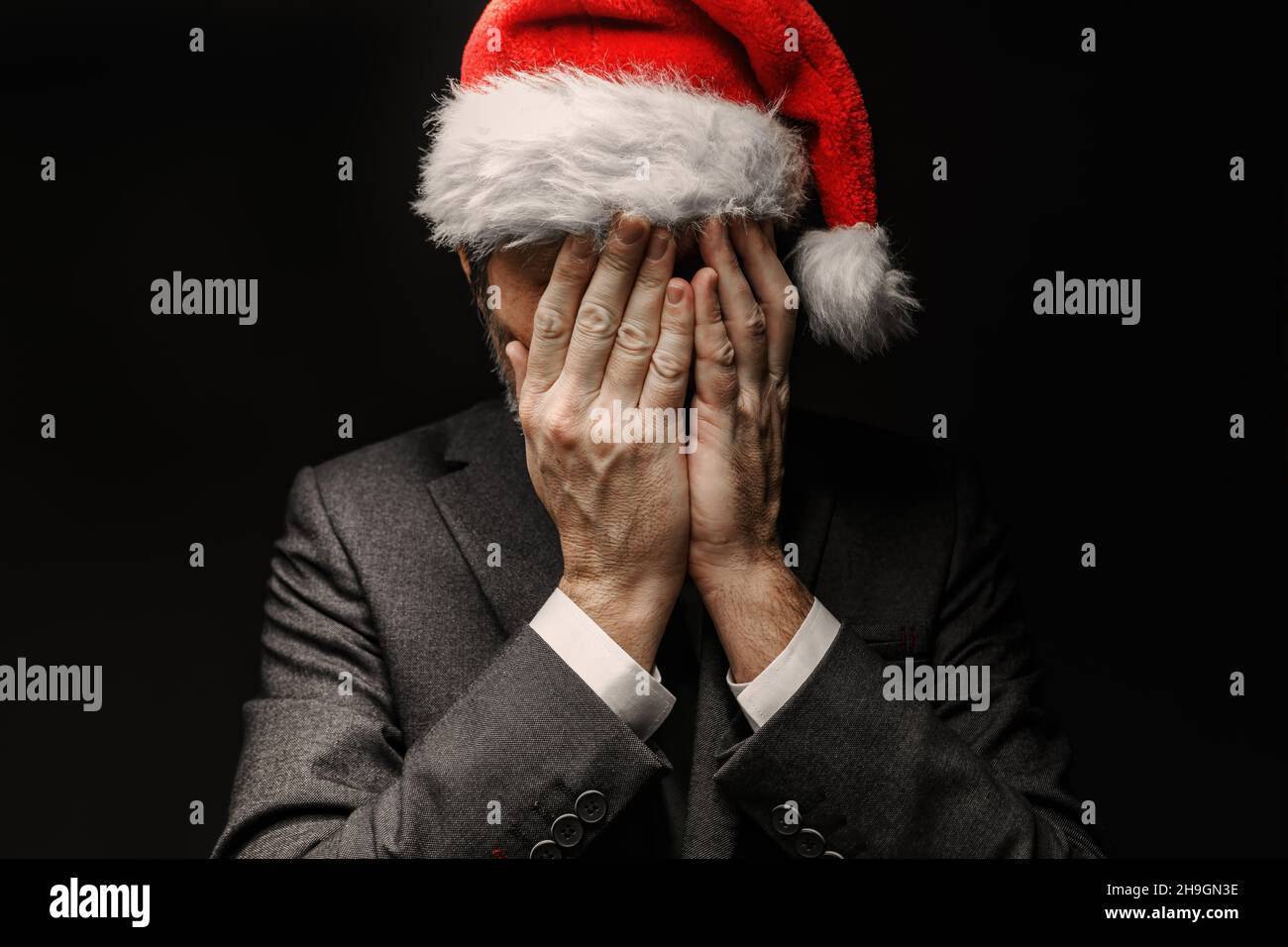 Very sad businessman with Santa Claus hat is crying with hands covering face, low key portrait with selective focus Stock Photo