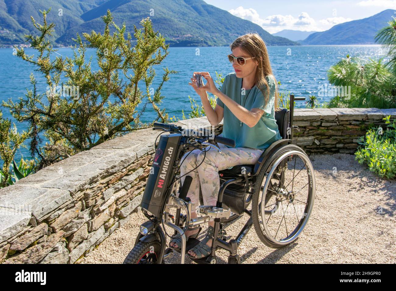 Woman in wheelchair at the lake side taking a picture of the scenery - junge Frau im Rollstuhl fotografiert am Seeufer die Landschaft - Brissago Stock Photo