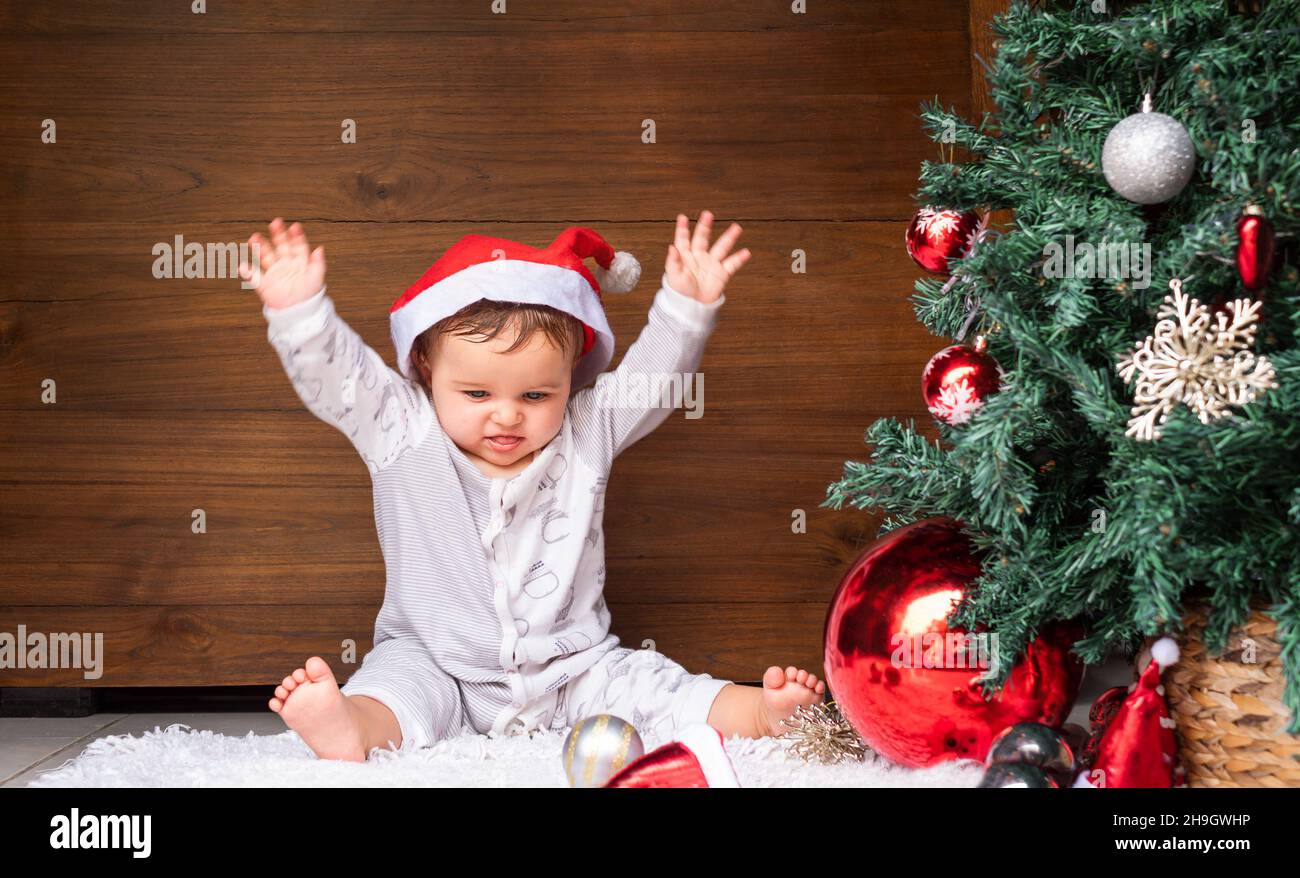 the baby sits on the floor near the tree and happily raises her hands up Stock Photo