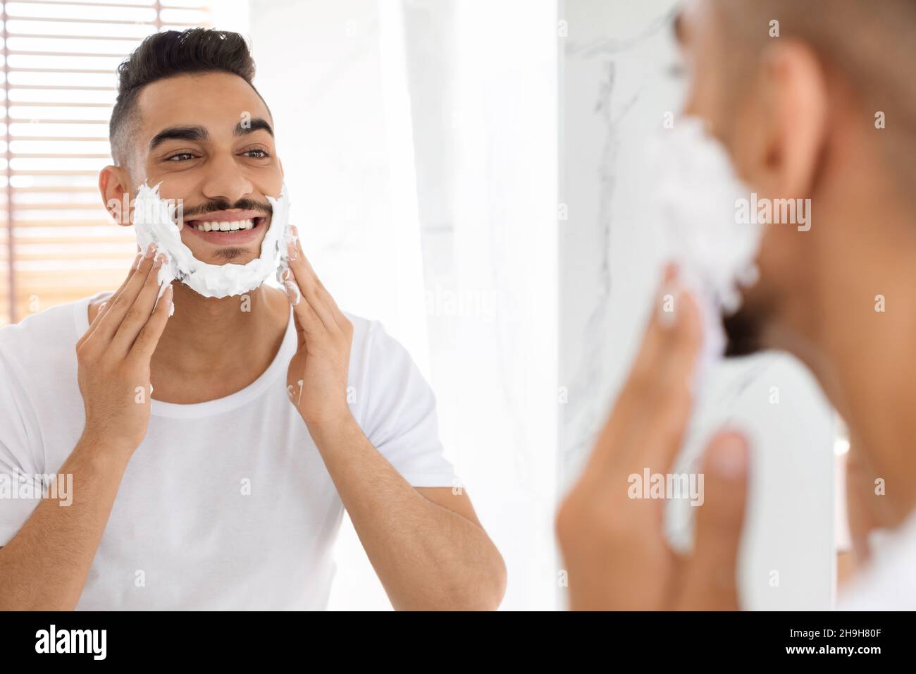Handsome Arab Man Applying Shaving Foam On Face Near Mirror In Bathroom