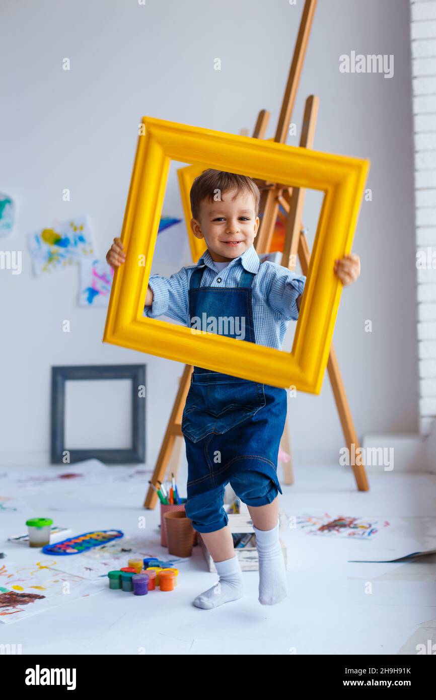 Cute, happy, white boy in blue shirt and jeans smiling and looking through yellow frame. Little child having fun in artist studio. Concept of early ch Stock Photo