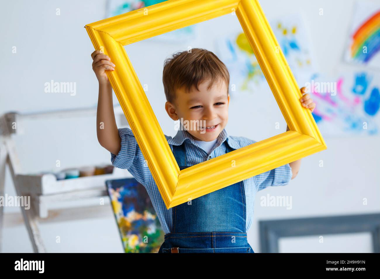 Cute, happy, white boy in blue shirt and jeans smiling and looking through yellow frame. Little child having fun in artist studio. Concept of early ch Stock Photo