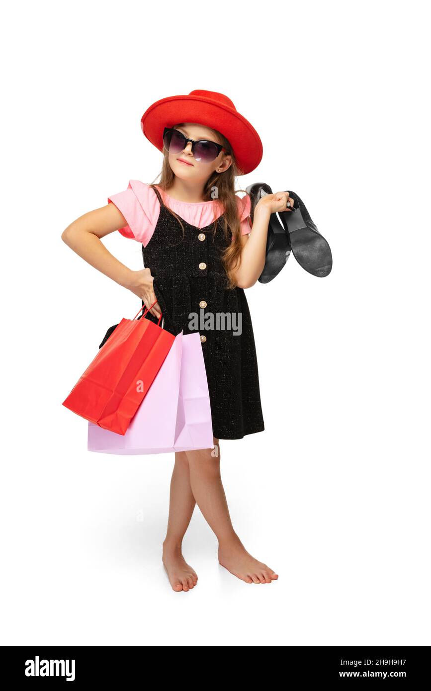 Stylish pretty little girl, charming kid standing with multicolored shopping packages and talking on phone isolated over white background. Stock Photo