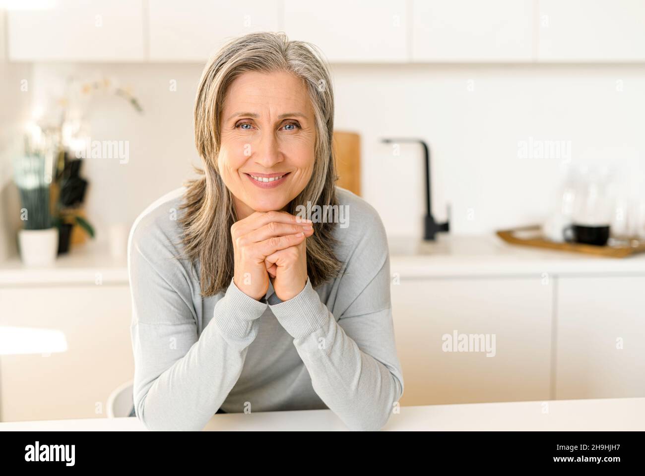 Joyful senior mature full of energy woman in casual blue pullover in the modern kitchen, gray-haired lady sitting at counter top resting chin on hands, middle-aged lady posing and looking at camera Stock Photo