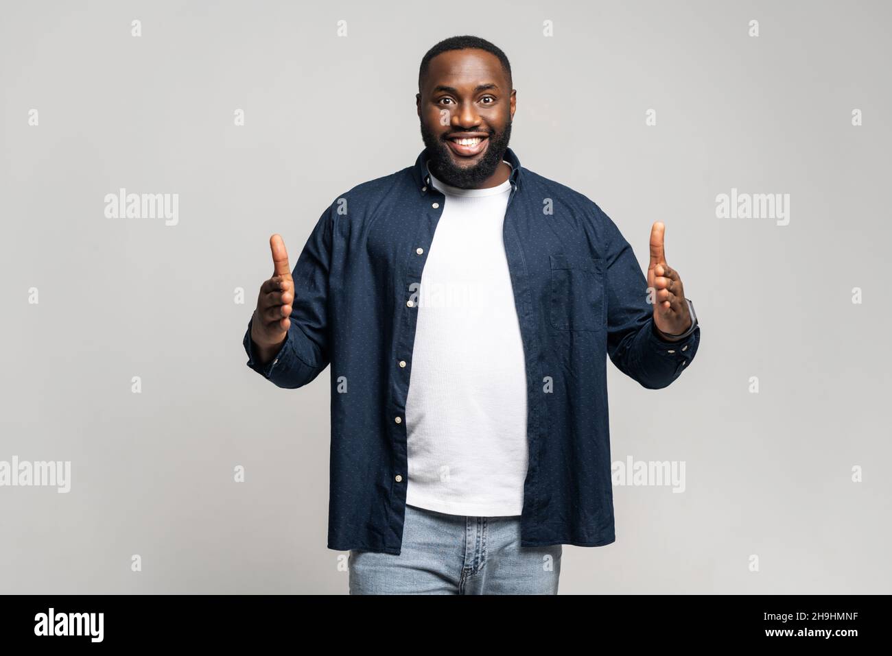 Positive African man with beard looks at camera, holding two hands in front of him showing the size on gray isolated background in studio, wearing casual shirt, smiling joyfully, presenting something Stock Photo