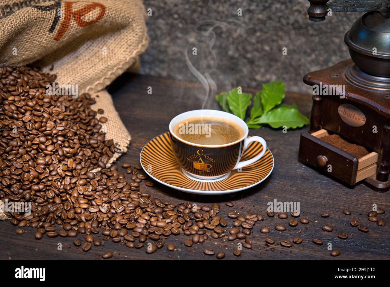 Kaffee Komposition mit dampfender Kaffeetasse, Kaffeebohnen in Kaffeesack, historischer Kaffeemühle und Kaffeeblätter auf einem Holztisch Stock Photo