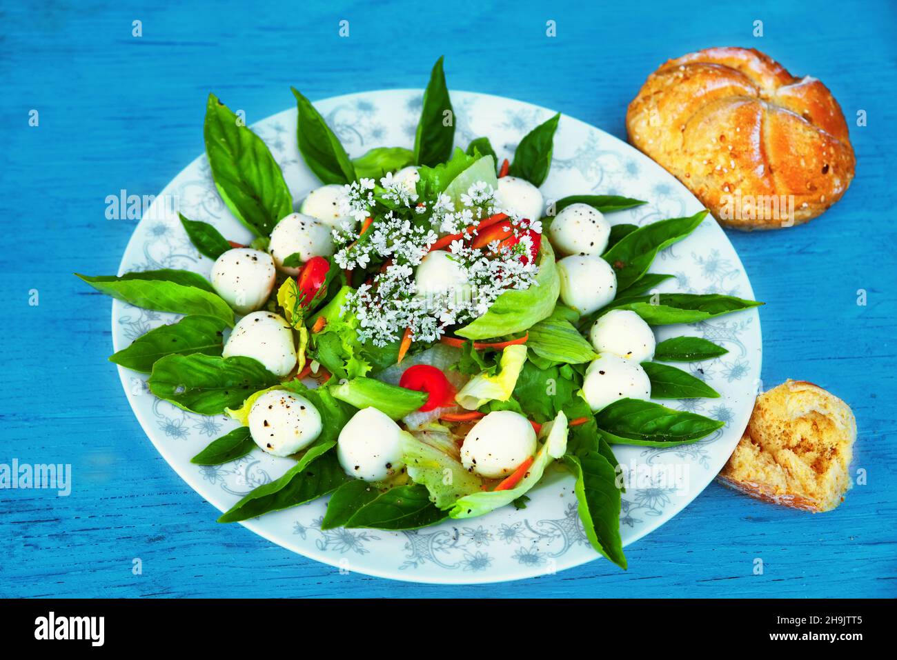 Plate with salad arranged in circle decorated with white tiny flower on blue wooden table. Mini mozarella ball, green basil leaf, olive oil and red pe Stock Photo