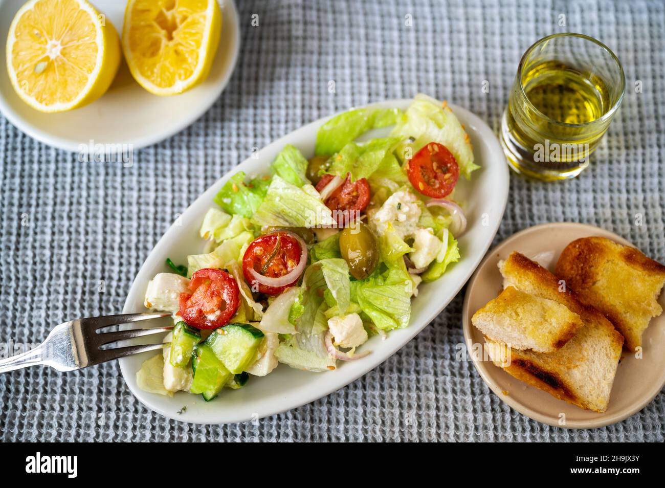 Plate with fresh salad (tomato,lettuce,cheese,cucumber,onion,olive), pastry, lemon and glass of olive oil. Stock Photo