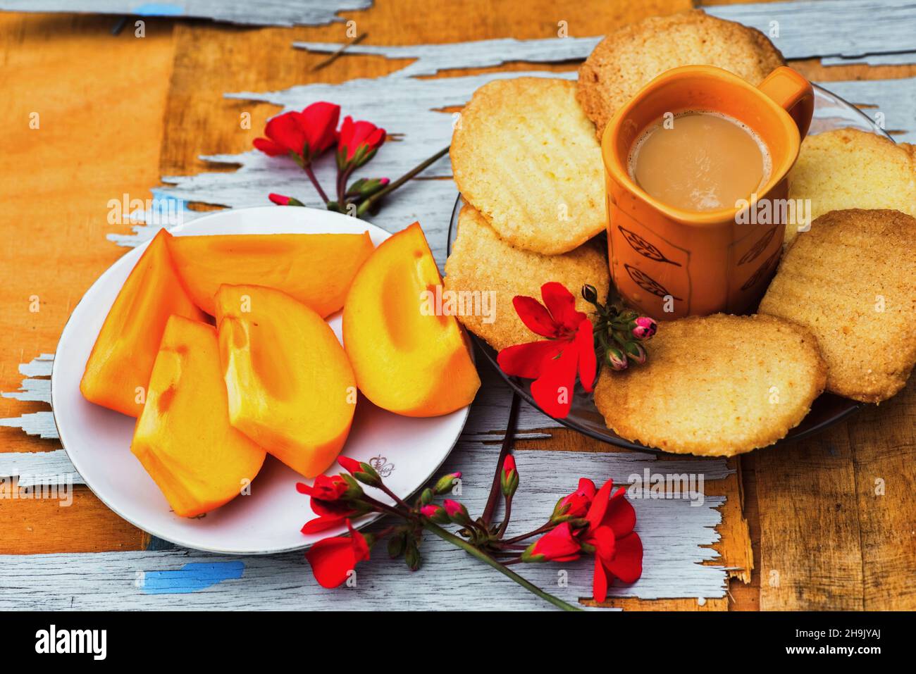 Sliced kaki fruit on plate, butter cookie, orange cup of coffee and red flower on scratched wooden table. Sweet food arrangement orange color. Stock Photo