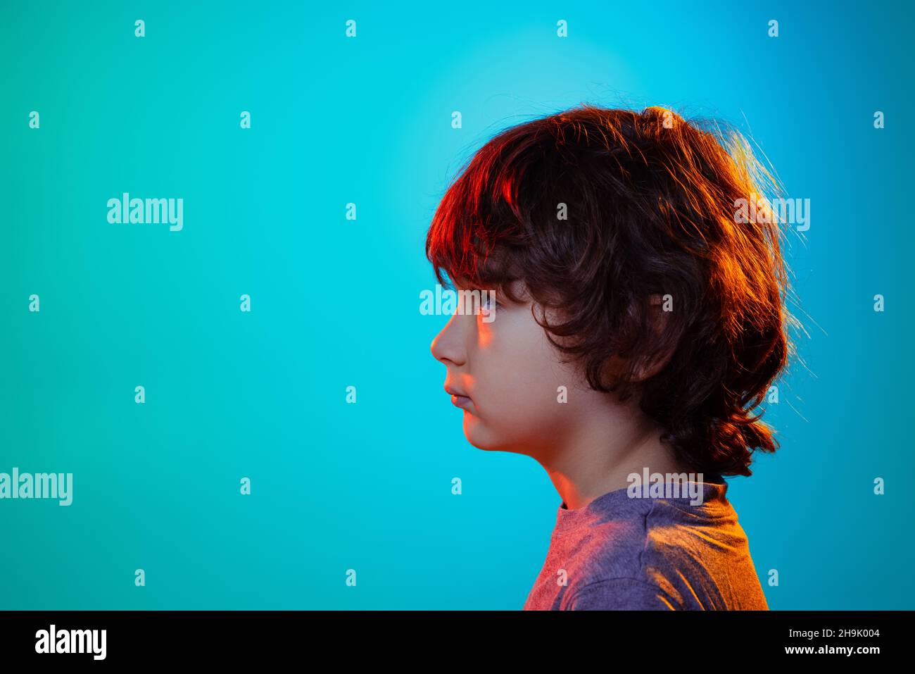 Profile view of little boy, curly kid posing isolated on blue studio background in neon light, filter. Stock Photo