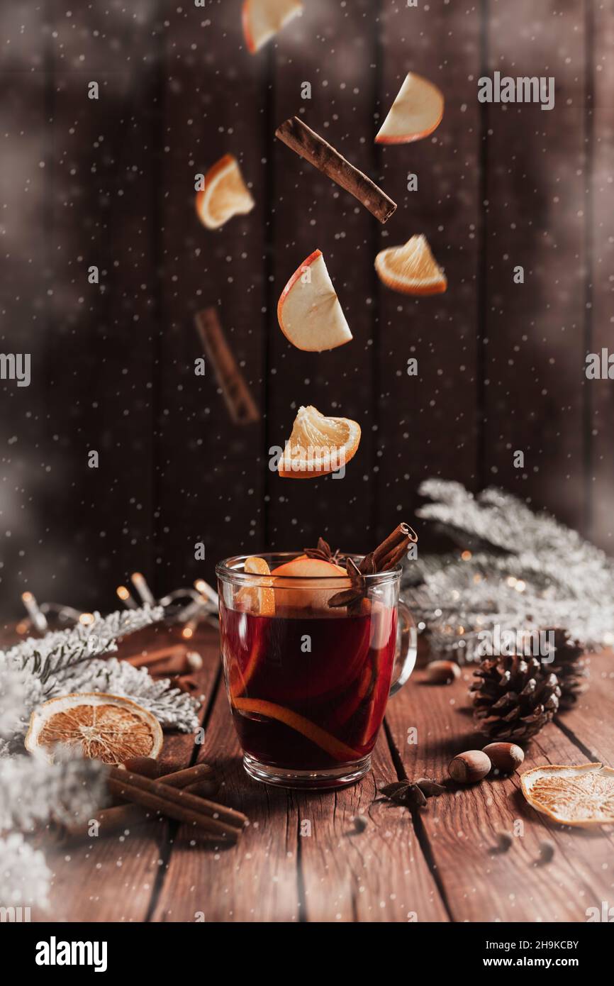 Christmas glass cup or mug with Mulled Wine on wooden table and background with levitating cinnamon stick, dried or sublimated orange, apple slices, anise, spices, hazelnuts, decoration light garlands Stock Photo