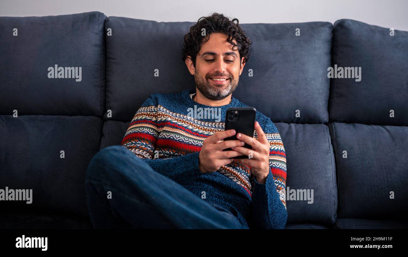 Happy handsome man smiling while using his smart phone and sitting on the couch at home. Confident bearded man relaxing and typing on smartphone at so Stock Photo