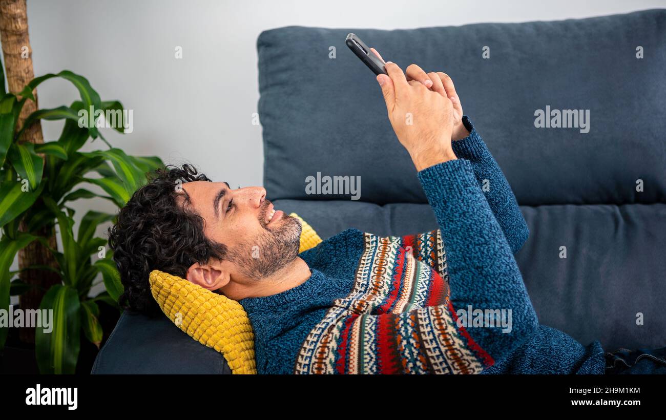Portrait of happy caucasian man smiling while using smartphone and lying down on couch at beautiful home interior. Caucasian man relaxing with phone d Stock Photo