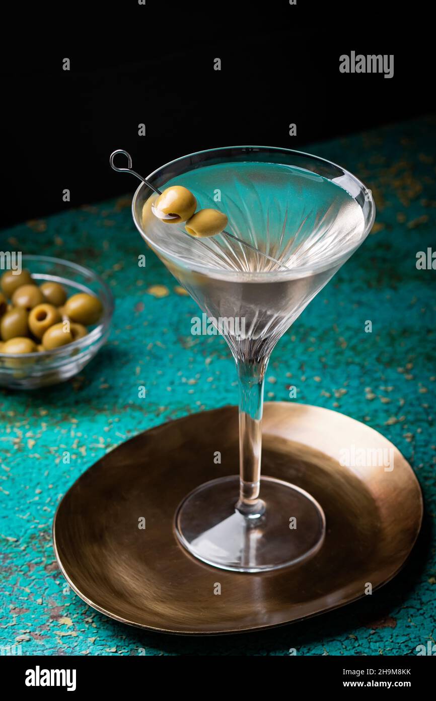 Dirty martini cocktail closeup in back light with olives garnish on green textured table closeup with plate with olives in background Stock Photo
