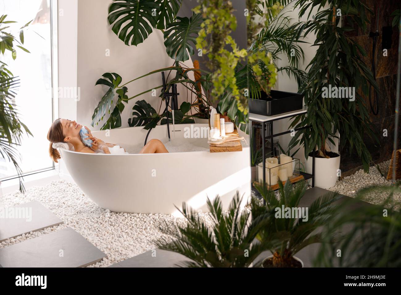 Relaxed young woman bathing in modern bathroom interior decorated with tropical plants Stock Photo