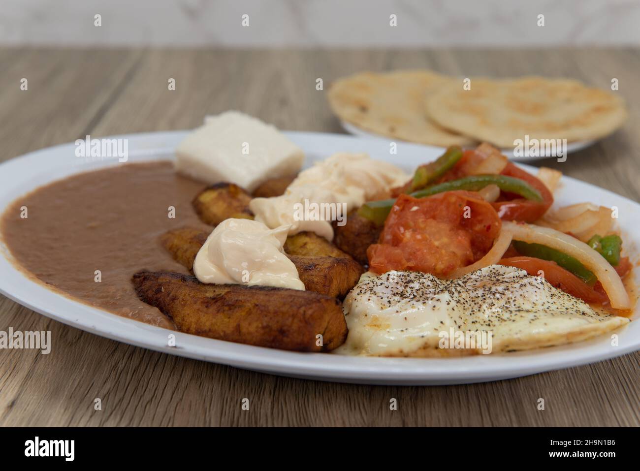 Fried plantains on a plate with refried beans, stir fried vegetables, and eggs served sunny side up with pupusa bread for a Latino meal. Stock Photo