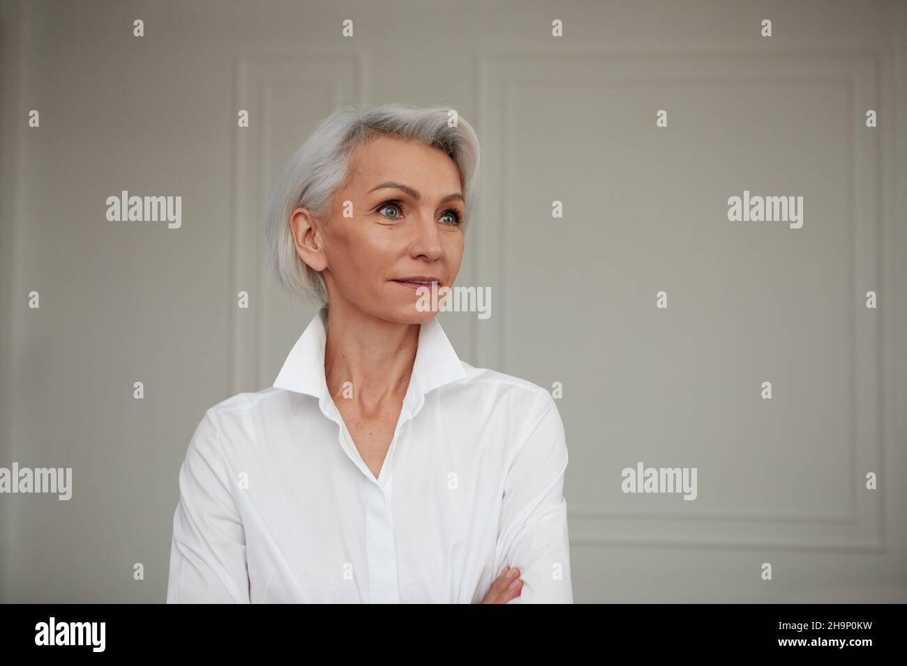 Portrait of an adult elderly self-confident woman with white hair looking awey into distance in white shirt hands crossed background wall Stock Photo