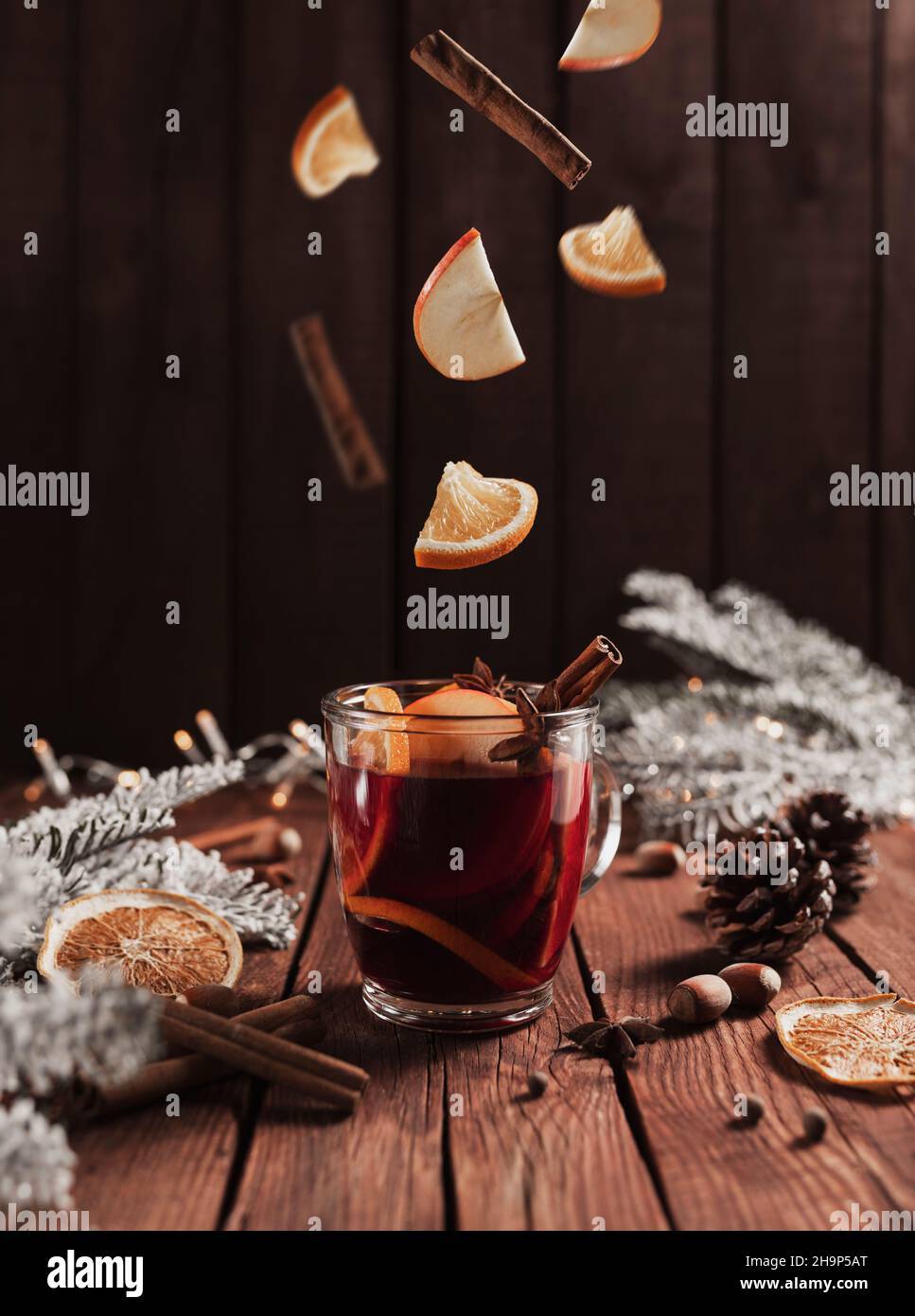 Christmas glass cup or mug with Mulled Wine on wooden table and background with levitating cinnamon stick, dried or sublimated orange, apple slices, present gift boxes, garlands, decoration lights Stock Photo