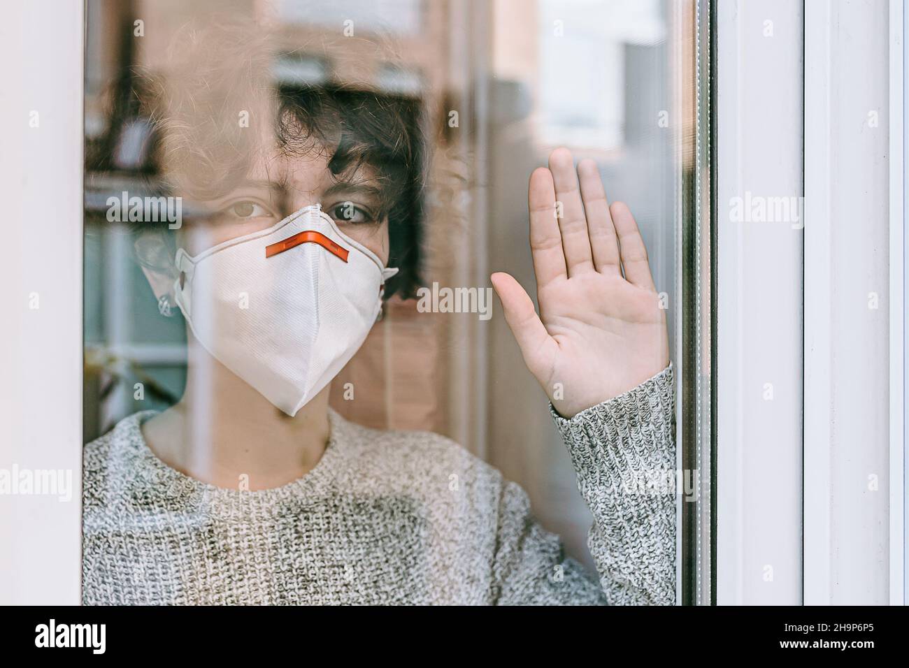 young teenage girl with brown curly hair wearing protective mask looks out window. quarantine and self-isolation for students and schoolchildren. Stock Photo