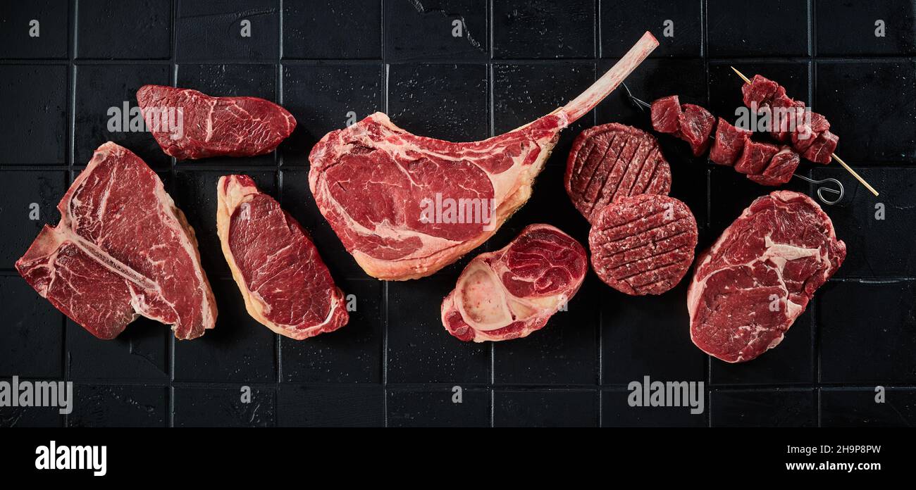 Top view of various uncooked beef steaks including t bone tomahawk and sirloins steaks near burger cuttles and shashlik on black table Stock Photo