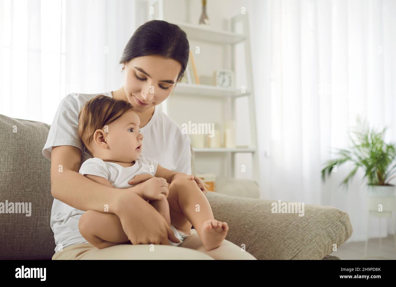 Young mother holding her cute baby on her lap while sitting on couch in the living room Stock Photo