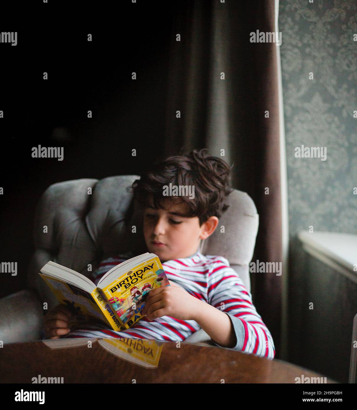 9 years old boy in pyjamas reading book on a chair next to a window Stock Photo